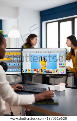 Black retoucher woman analysing images on pc with two monitors and retouching digital assets with stylus pencil. African project designer using photo editing software sitting in creative agency office
