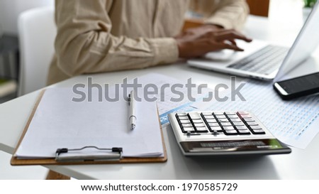 Clipboard and calculator on the desk with people working with laptops in the background.