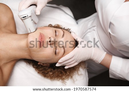 Spa salon and hardware cosmetology. SMAS non surgical ultrasound facelift procedure carried out by beautician dermatologist in beauty clinic. Ultrasonic device produce collagen on client face skin Royalty-Free Stock Photo #1970587574