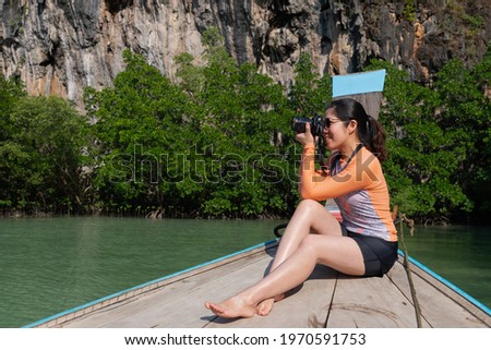 young asian woman weraing swimsuit using camera to take photo while travel on long tail boat on summer vacation