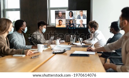 Videoconferencing, Virtual Meeting. Diverse group of workers in masks making online videocall with colleagues, sitting at table in boardroom, looking at screen, talking on web with remote employees Royalty-Free Stock Photo #1970614076