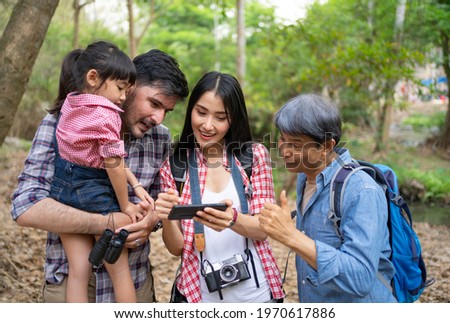 Happy family, Caucacian father, Asian mother, daughter and grandfather standing among forest trees looking at pictures just taken on smartphone happily while backpack hiking in shady forest on holiday