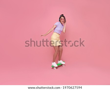 Young asian teenage girl playing on skateboard with wearing wireless headphones listening to music on pink background.