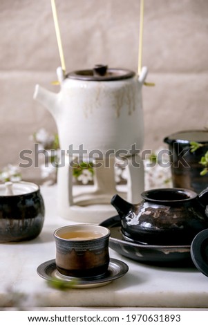 White and black craft handmade ceramic teapot with black ceramic mugs, katakuchi jug and blossoming cherry tree as decoration standing on marble table. Traditional Japanese tea ceremony. Teadrinking