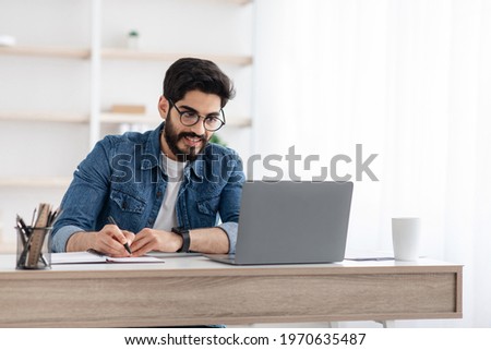 Arab businessman taking notes while working on laptop computer at home office, empty space. Freelancer noting down information from internet, planning working schedule, sitting at workplace