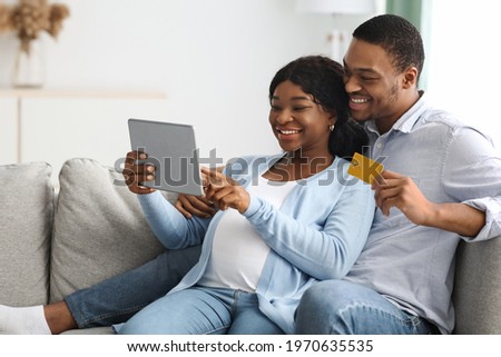 Happy black pregnant couple shopping online from home, using credit card and digital tablet, copy space. Positive african american expecting family purchasing on Internet, buying baby clothes