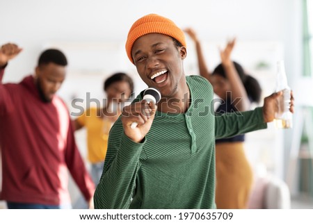 Positive african american young man with bottle of beer singing karaoke while having home party with friends, holding microphone and singing songs, selective focus, blurred background Royalty-Free Stock Photo #1970635709