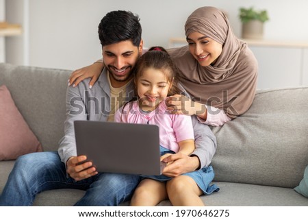 Precious time with family. Happy muslim parents and cute little girl using laptop together, watching photos or movie, browsing internet, sitting on sofa in living room