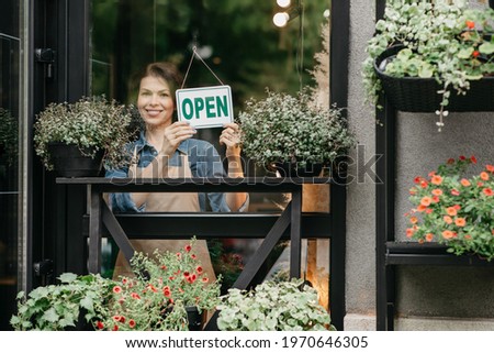 Be protected, after coronavirus rules, opening of store, cafe and salon in city. Happy young pretty woman in apron turns sign with inscription open, on glass door in flower studio with plants