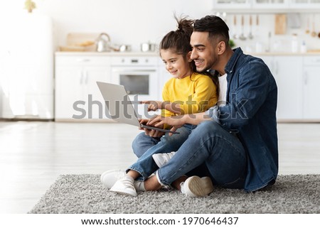Happy Arab Man With His Little Daughter Using Laptop Together At Home, Middle Eastern Dad And Female Child Using Computer For Online Shopping Or Browsing Internet While Relaxing On Floor In Kitchen Royalty-Free Stock Photo #1970646437