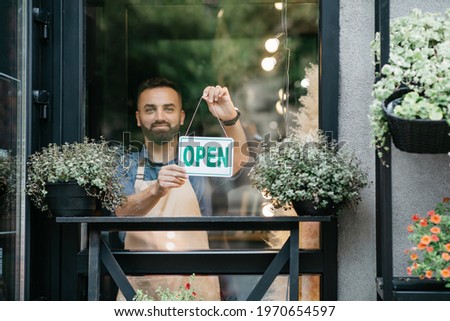 New rules for store opening after covid-19 quarantine and positive emotions of owner. Happy young attractive bearded guy in apron turns sign with inscription is open on door in flower studio, outdoor