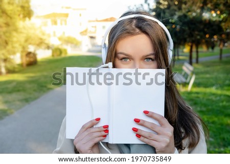 Young woman in white electronic headphones cover her face with white book in nature outdoor. Listening online audiobook. Enjoying music or leisure podcast. Storytelling. Text space