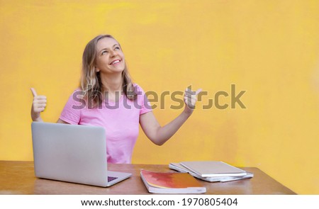 Woman sitting at table with laptop and showing thumbs up on yellow background