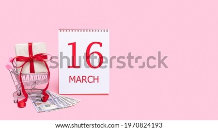 16th day of march. A gift box in a shopping trolley, dollars and a calendar with the date of 16 march on a pink background. Spring month, day of the year concept.