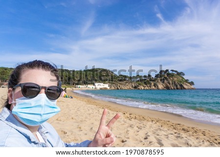 Close-up of a woman with a protective mask waving at the camera in front of the sea