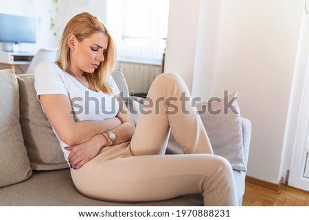 Young sick woman with hands holding pressing her crotch lower abdomen. Medical or gynecological problems, healthcare concept. Young woman suffering from abdominal pain while sitting on sofa at home Royalty-Free Stock Photo #1970888231