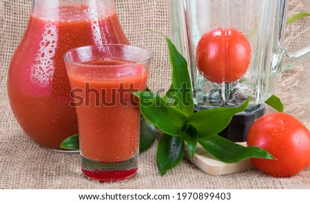 Tomato juice in glass pitcher under water drops, blender, greens, two raw tomatoes on burlap background