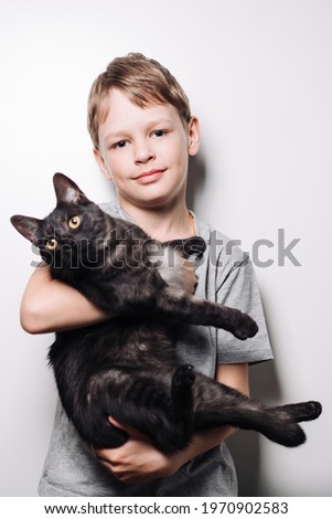 Boy with blond hair and in a gray t-shirt hugs a cute dark cat on a white background