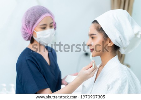Asian female beautician in blue clothes, medical hat, and face mask applying facial peeling masks on beautiful Asian female client's face who smiling sitting on bed in a beauty clinic's treatment room