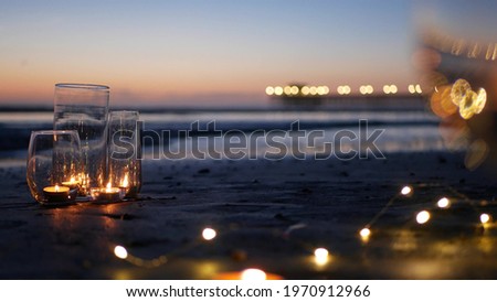 Candle flame lights in glass, romantic beach date, California ocean waves, sea water. Candlelight seamless looped cinemagraph. Wineglass on sand, garland in twilight dusk. Illuminated pier reflection. Royalty-Free Stock Photo #1970912966
