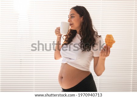 Beautiful pregnant woman holding croissant and cup of coffee in her hands during morning breakfast. Concept of good health and positive attitude while expecting baby. Copy space.