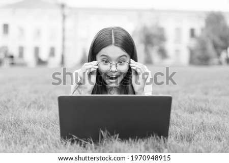 Surprised school kid in glasses playing online game on laptop relaxing on green grass in park, teenage free time