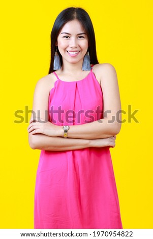 Asian beautiful cheerful woman with long hair wearing pink dress, fashionable earring, wrist watch, happily smiling with confidence, crossing arms, looking at camera with isolated yellow background