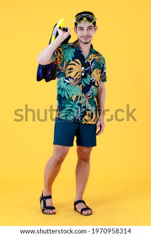 Attractive young Asian man smiles and holds swim fins and snorkel mask. Full body studio portrait on yellow background. Summer holiday travel preparation concept