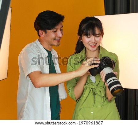 Male model smiling, viewing his picture on DSLR camera screen after taken by young female photographer in the modern studio with orange background and they are very pleased with the image