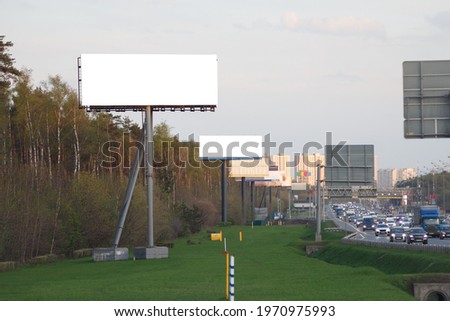 Tall empty billboard in the woods by a busy highway against the backdrop of a large city in summer.