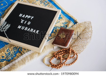 Letter board says Happy Eid Mubarak on the prayer mat with holy book Al Quran and prayer beads. There is Arabic letter which means the holy book