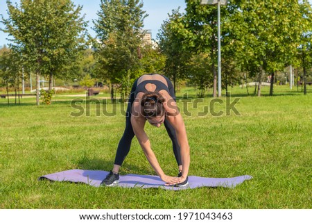 Muscular middle aged caucasian woman makes stretching in city park on green grass. Tilt down exercises. Black sportswear. Stretching training. Healthy lifestyle. Outdoor sport theme.