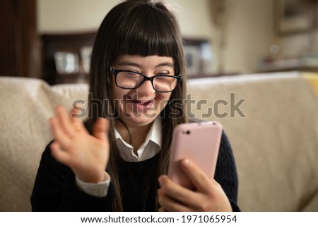Cinematic shot of happy teen girl with down syndrome making video call with smartphone to friends or family at home. Concept of technology, disability, media, friendship, new generation. Royalty-Free Stock Photo #1971065954