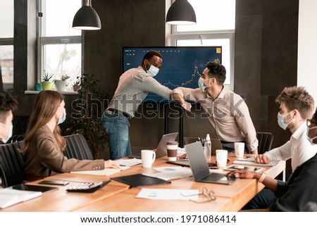 Stop Spreading Virus Concept. Diverse colleagues wearing protective face masks greeting each other and bumping elbows at office. Workers avoid a handshake during meeting, keeping social distance Royalty-Free Stock Photo #1971086414