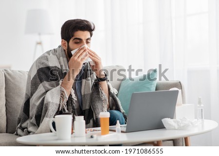 Ill arab man covered in warm blanket looking at laptop screen and sneezing nose in paper tissue, sitting on sofa at home, empty space. Sinusitis illness, cold and influenza symptoms Royalty-Free Stock Photo #1971086555