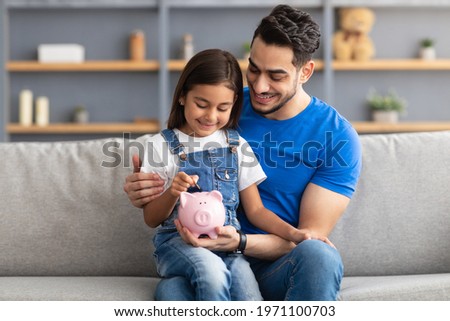 Financial Education For Children Concept. Portrait of smiling little girl putting coin in pink piggy bank, sitting on dad's lap on the couch at home, man teaching his daughter to invest Royalty-Free Stock Photo #1971100703