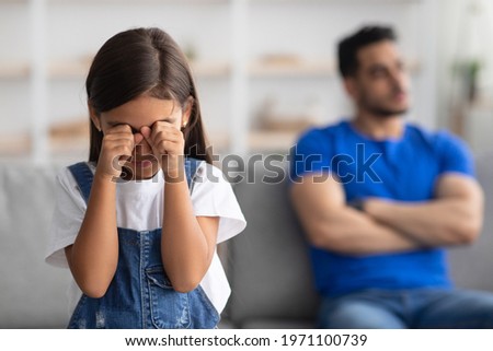 Family Relationship Problems. Selective focus on offended crying little kid girl wiping tears, young father sitting separate on couch in the blurred background, ignoring daughter. Generations gap Royalty-Free Stock Photo #1971100739