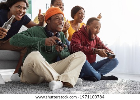 Happy african american fellows having fun at home, drinking beer, emotional black guys with joysticks playing video games, gesturing and screaming, enjoying time together, copy space