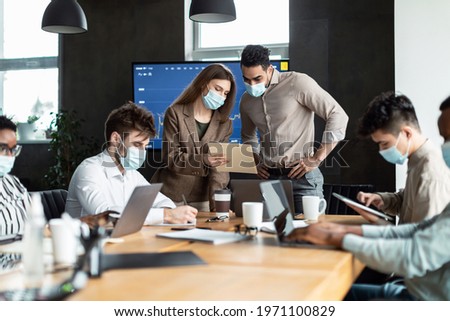 Teamwork And Corporate Meeting Concept. Businesspeople In Medical Face Mask Working At Office, Having Breefing With Coworkers, Young Man Standing At Table In Boardroom, Secretary Showing Documentarion