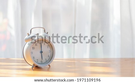 White alarm clock on desk with beautiful curtain for background. Concept of light, hope and the beginning of new day.