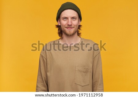 Cool looking male, handsome bearded guy with blond hair. Wearing green beanie and beige sweater. Watching at the camera with confident smile, isolated over yellow background