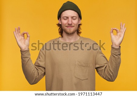 Young man, calm looking guy with blond hair, beard and mustache. Wearing green beanie and beige sweater. Peacefully meditating with closed eyes. Stand isolated over yellow background