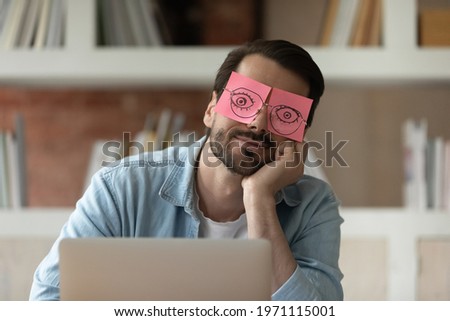 Close up tired businessman with stickers on face sleeping, drawn eyes on adhesive papers, sitting at work desk in office, unproductive lazy young male dozing, working on difficult project, fatigue Royalty-Free Stock Photo #1971115001
