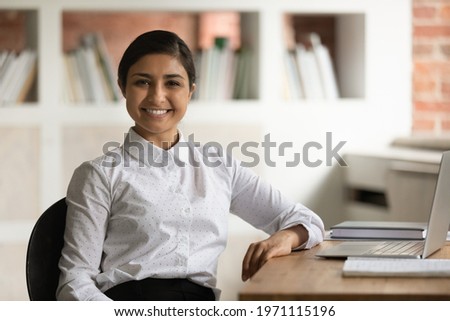Head shot portrait smiling attractive Indian businesswoman sitting at work desk in office, confident happy young woman looking at camera, posing for corporate photo, motivated student or employee Royalty-Free Stock Photo #1971115196