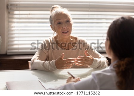 Visiting doc. Aged female pensioner sit by table at doctor office talking sharing complaints asking medical worker questions. Old lady patient describe disease symptoms to woman physician on checkup Royalty-Free Stock Photo #1971115262