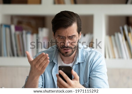 Close up unhappy irritated man in glasses looking at phone screen, worried businessman reading bad news in message, confused young male having problem with broken or discharged device, data loss Royalty-Free Stock Photo #1971115292