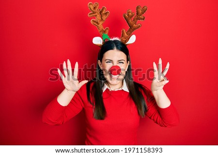 Young hispanic woman wearing deer christmas hat and red nose showing and pointing up with fingers number eight while smiling confident and happy. 