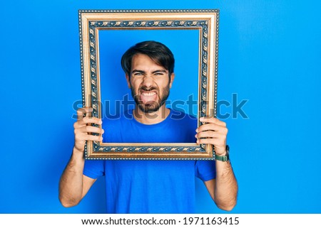 Young hispanic man holding empty frame sticking tongue out happy with funny expression. 