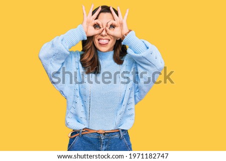 Young brunette woman wearing casual winter sweater doing ok gesture like binoculars sticking tongue out, eyes looking through fingers. crazy expression. 