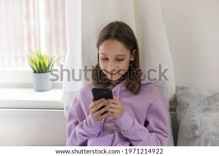 Happy young teenager girl in comfortable hoodie looking at telephone screen, involved in playing video game or communicating distantly in social network, modern tech addiction and Z generation people.